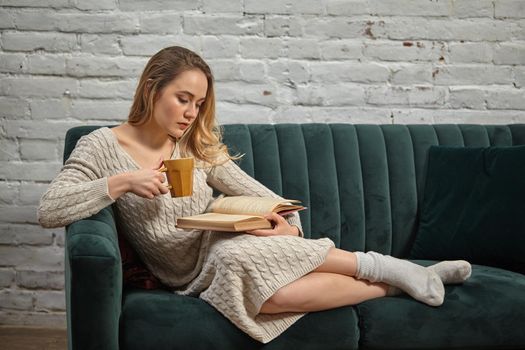 Blonde model blogger in beige knitted dress and socks is reclining on gray sofa, holding brown cup and reading book against white brick wall. Close-up