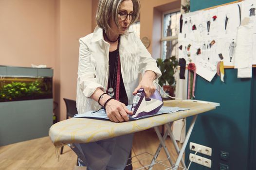 Inspired mature European white-haired woman fashion designer tailor using steam iron to press blue shirt from new collection on an ironing board in a clothing design and tailoring atelier
