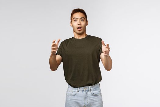 Portrait of shocked young male, taiwanese guy catching product that person throwing at him, raising hands up and look intense and stressed, gasping startled, standing grey background