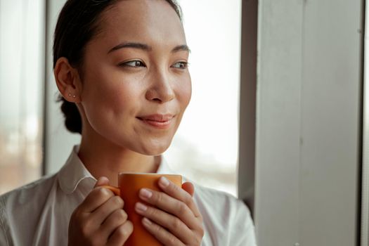 Smiling Asian business woman enjoying coffee standing near office windows