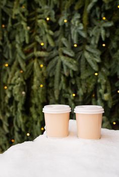 Two paper coffee cups outdoors in the snow against the backdrop of a beautiful green nobilis wall, coffee to go concept. Walks in the open air.