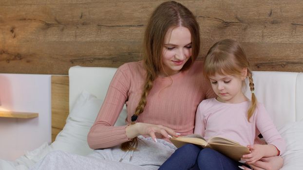 Young mother reading goodnight story book fairytale to child daughter kid girl in night bedroom