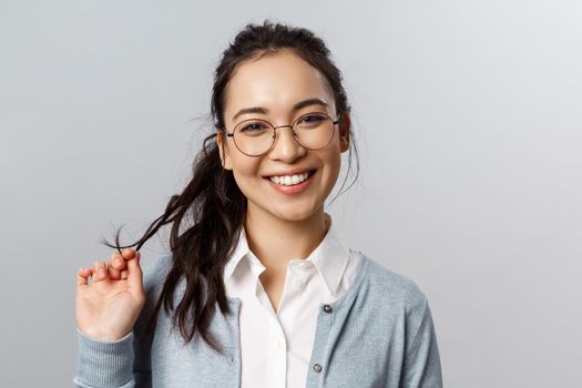 Close-up portrait of flirty, enthusiastic brunette asian female in glasses, beaming smile, roll strand of hair between fingers as coquettish talking to person she likes, standing grey background