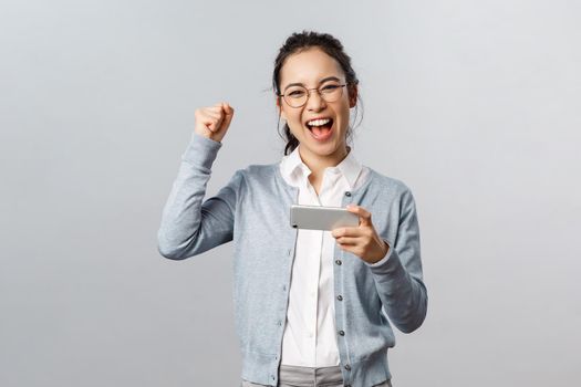 Technology, online and mobile lifestyle concept. Excited, happy triumphing asian woman winning prize or beat score in smartphone game, holding phone, raise hand up celebrating victory