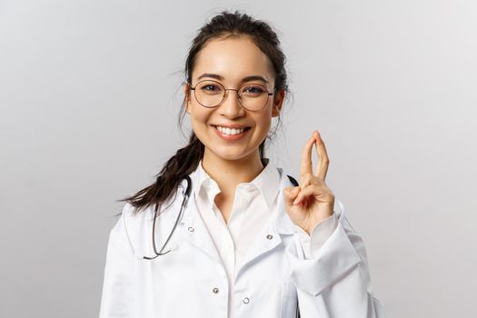 Close-up portrait of enthusiastic, smiling attractive asian female doctor, therapist check-up child in hospital, showing few tricks to cheer patient, standing upbeat grey background