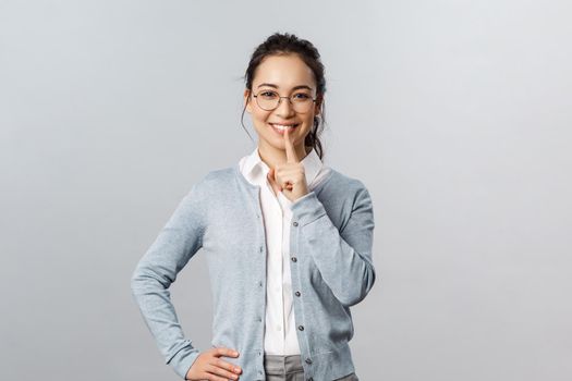 Education, studying and online classes concept. Charming young asian girl in glasses asking polite to keep quiet, shush with smile, hold finger over mouth, be silent please, hold it a secret
