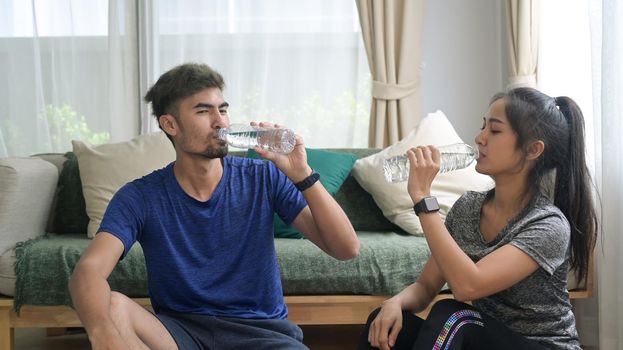 Young couple in sports clothes is drinking water, resting on the floor after workout