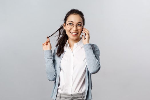 Office lifestyle, business and people concept. Talkative cute and silly female meployee, asian girl roll strand of hair on finger, look away dreamy and smiling, talking on phone gossiping
