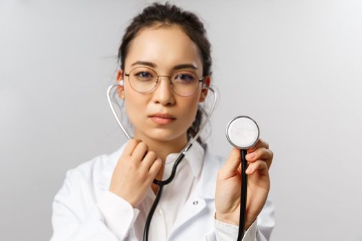 Covid19, coronavirus, healthcare and doctors concept. Portrait of serious-looking female doctor during check-up of patient in ER, hospital, wearing white coat, listening to lungs with stethoscope