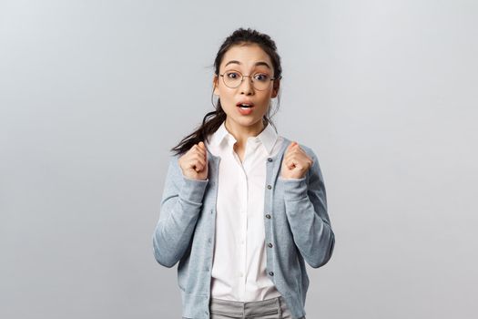 Portrait of excited and tempting cute asian girl in glasses, awaiting with thrill and interest for new season of favorite tv show coming up, looking ancitipation and amusement, stand grey background
