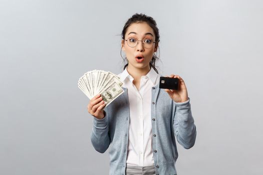 Business, finance and investment concept. Amazed curious asian female in glasses, look impressed and surprised, holding dollars cash and credit card, decide how better keep money safe