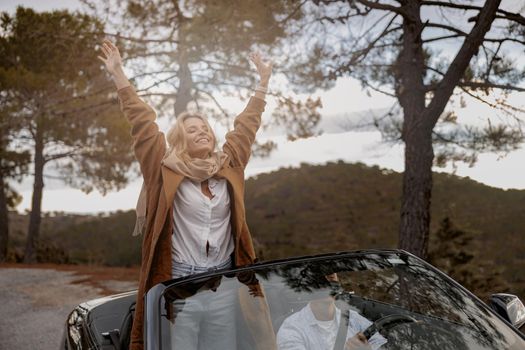 Pretty young smiled woman stands on front seat in convertible car with her hands raised up