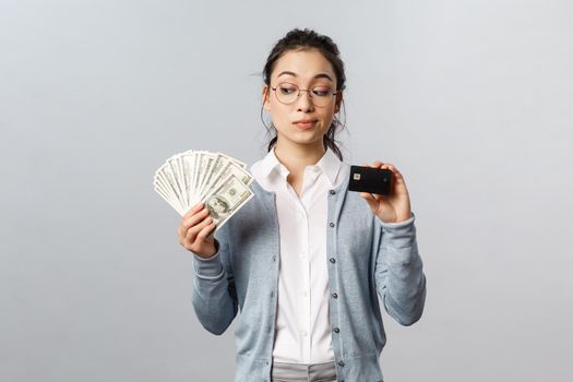 Business, finance and investment concept. Thoughtful indecisive cute asian girl look tempting at cash dollars, holding money and credit card, shopping, think where invest, grey background