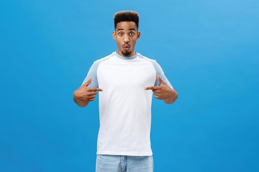 Impressed and amazed good-looking young dark-skinned male model in casual t-shirt folding lips in intrigue and curiosity popping eyes at camera excited while pointing at chest over blue wall