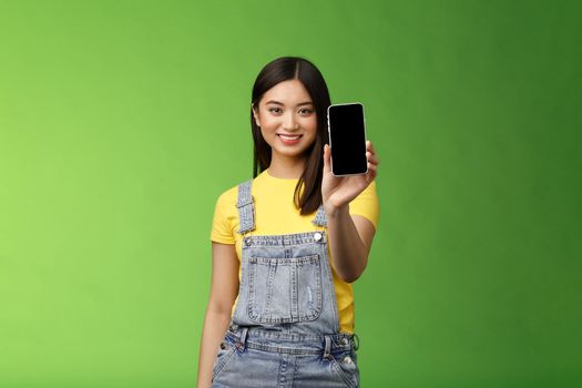 Confident assertive good-looking female asian introduce smartphone app, extend hand hold telephone arm, smiling delighted, recommend social profile, bragging own blog, green background