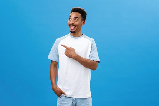 Studio shot of friendly joyful and emotive handsome dark-skinned bearded guy with afro haircut pointing and looking left with excited thrilled face smiling broadly posing over blue background