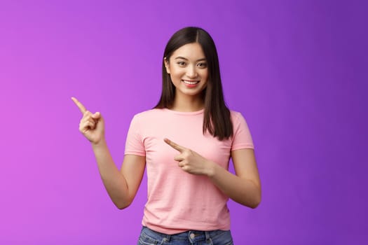 Friendly cute pleasant asian woman showing way, indicating interesting promo, pointing left smiling broadly, introduce advertisement product, stand purple background cheerful