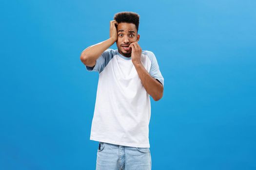 Insecure anxious guy getting scared of consequences. Portrait of frightened panicking handsome young african american male biting fingernails gazing worried at camera posing over blue background