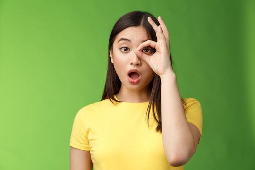 Amazed speechless cute asian young girl drop jaw astonished, look through ok circle sign, drop jaw speechless, impressed look camera, check out incredible promo, stand green background