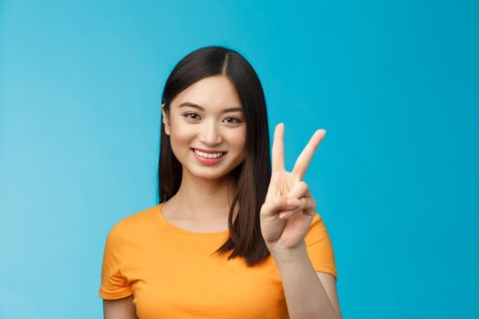 Cheerful friendly asian woman in yellow t-shirt smiling broadly, show number two, twice, win second place, smiling joyfully, make take-away order, book sits, stand blue background