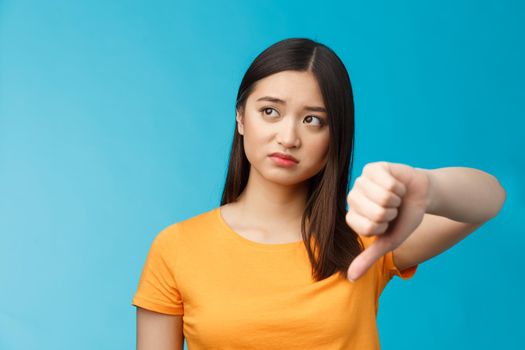 Disappointed female asian visitor give negative feedback, look away disturbed and upset, show thumb down dislike sign, judging bad thing, poor quality, stand blue background