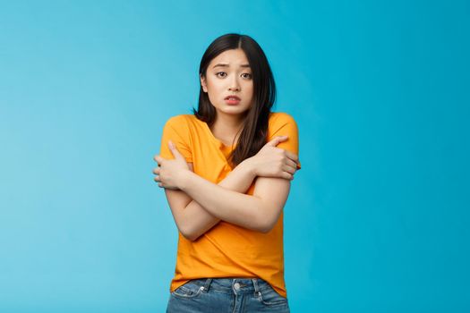 Girl feeling uncomfortable walking light yellow t-shirt, hugging herself trembling, shaking feeling cold, freezing windy weather, frowning grimacing discomfort, stand blue background