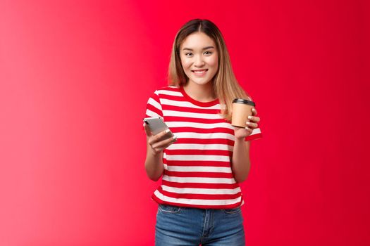 Cute blond asian university girl student stand lunch break enjoy coffee near campus, hold smartphone, messaging friend, waiting girlfriend park, order take-away drink smiling broadly, red background