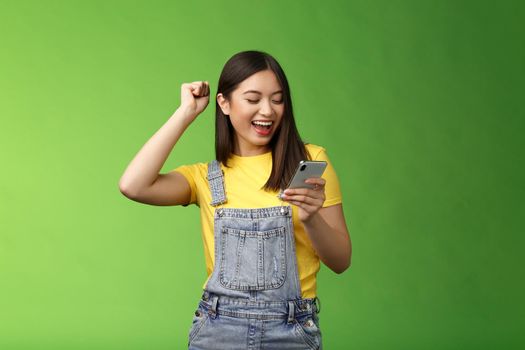 Triumphing cute joyful asian girl hold smartphone, fist pump celebrating good news, read good news message, look telephone screen smiling happily, achieve success got persmission party