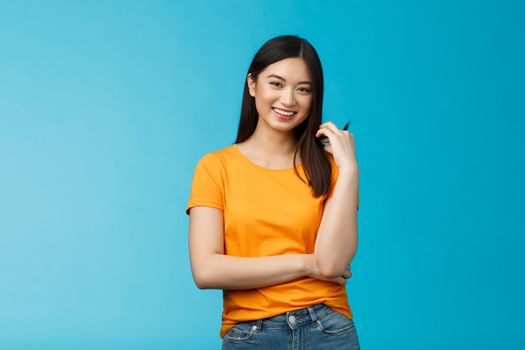 Cheerful asian girl with dark short haircut touching hair strand smiling lively, cross one arm chest, laughing, carefree talking casually friends, discuss university lifestyle, blue background