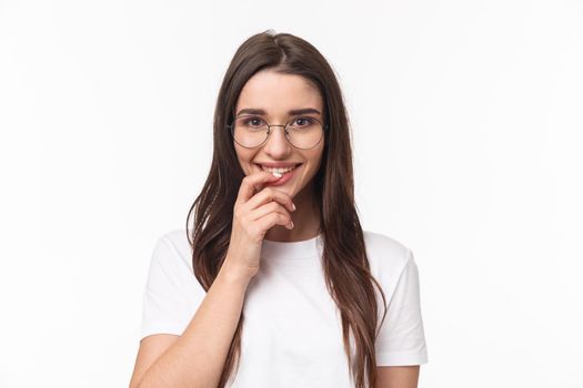 Close-up portrait of excited feminine, cute young woman in glasses, smiling and looking with temptation or desire, touching lip, want try something tasty, standing white background