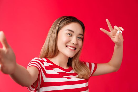 Friendly outgoing cute asian girl blond short hairstyle extend arm, hold smartphone camera, tilt head joyfully show peace victory sign, taking selfie photographing herself, stand red background