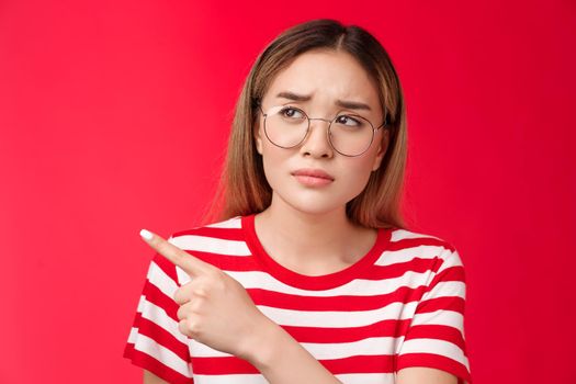 Disappointed doubtful frowning asian girl asking question reassure, looking pointing left displeased, unimpressed with strange place, dislike interiour sulking distressed, stand red background