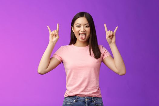 Rebellious amused asian girl enjoy rock-n-roll, attend awesome concert have fun, show tongue make heavy-metal gesture, express excitement and joy, stand purple background cheerful