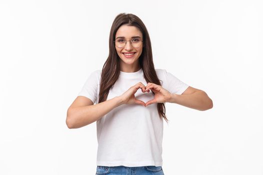 Portrait of caring and romantic lovely young woman in glasses, t-shirt, showing heart sign and smiling, express love care and sympathy, standing white background, open her heart to you