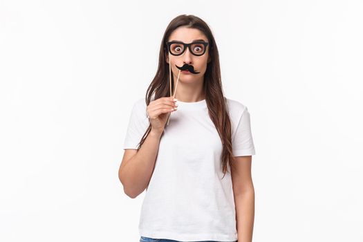 Entertainment, fun and holidays concept. Portrait of funny, playful and carefree girl holding carnaval mask sticks, fancy moustache and glasses, staring camera with popped eyes, white background