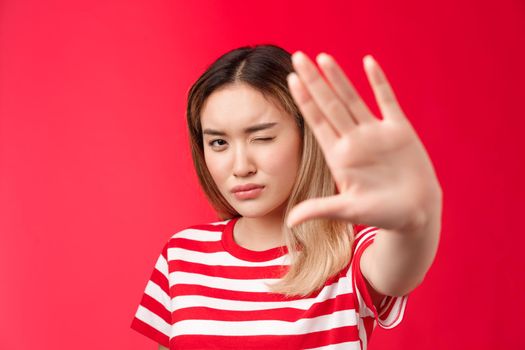 Close-up serious-looking determined asian blond woman pull hand forward show stop taboo sign, concentrating gaze on camera look with one eye, demanding stop, tell enough, say no, red background