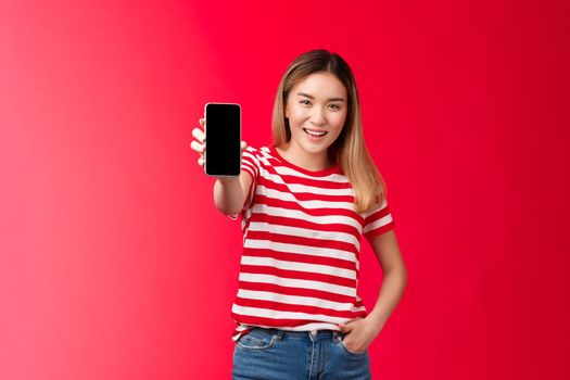 Check out my game score. Carefree good-looking blond asian woman extend arm showing smartphone screen smiling broadly stand relaxed introduce cool app, bragging bank account red background
