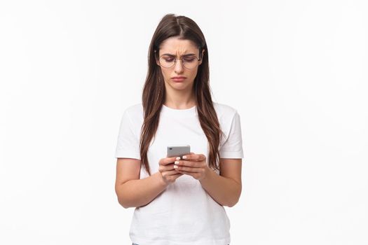 Communication, technology and lifestyle concept. Portrait of troubled and confused young woman reading strange message, frowning as looking mobile phone display, stand white background