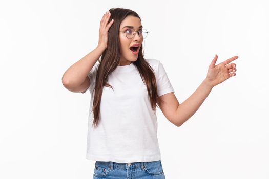 Waist-up portrait of surprised, astonished young brunette woman, 20s years, pointing and looking right, touch head cant believe she sees something so awesome, startled standing white background