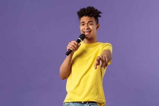 This song is for you. Portrait of romantic carefree hispanic man singing karaoke, pointing at camera as dedicate his performance, holding microphone, standing purple background