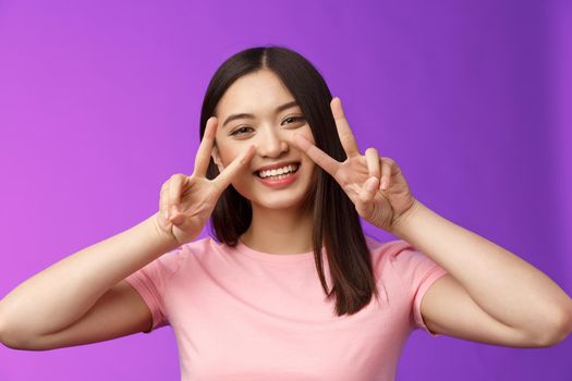 Close-up friendly positive outgoing nice asian girl show peace, victory signs cherish friendhsip stay optimistic, smiling broadly, having funny summer holidays, stand purple background