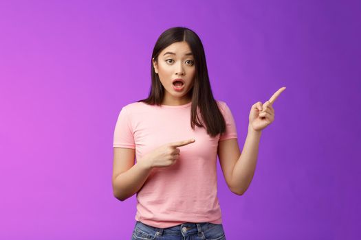 Excited alarmed cute asian female describe shocking scene pointing right, show index fingers sideways thrilling event, discuss amazing performance look camera worried, stand purple background