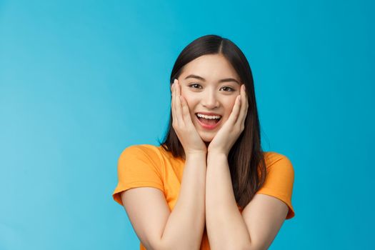 Flirty cute asian girl look surprised, express admiration and joy, touch cheeks, blushing coquettish smiling receive good news, stand blue background happily react amazed and excited