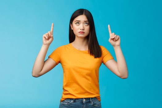 Upset gloomy cute asian young girl dark haircut frowning pointing looking up uneasy, feel regret cannot effort buy cool thing, stand blue background unsatisfied frustrated, wear yellow t-shirt