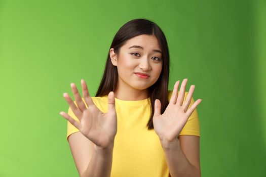 Modest cute insecure timid asian girl waving hands no gesture, step back, smirk, cringe aversion and dislike, refusing suspiciosu offer, rejecting proposal, stand green background