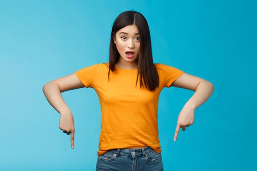 Impressed asian girl drop jaw shocked look camera surprised pointing down indicaring bottom advertisement, gasping open mouth astonished curious about interesting opportunity, blue background