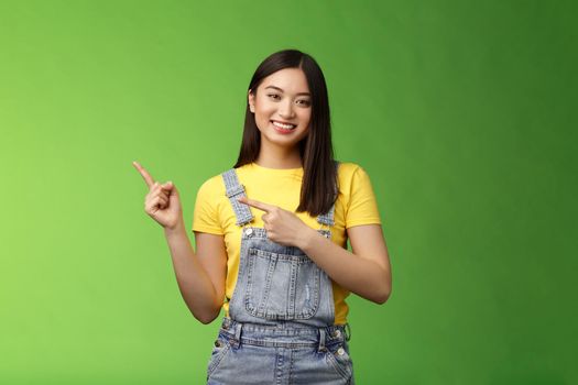 Lovely pretty tender asian woman showing way, tilt head flirty smiling, helpful pointing left, introduce plan, gladly present promo recommend cool new product, stand green background happy