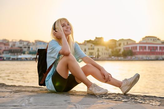 Happy teenage female tourist sitting on sea promenade of an old city in sunset