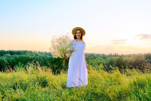 Beautiful woman in white dress hat with bouquet of flowers on meadow, in sunset