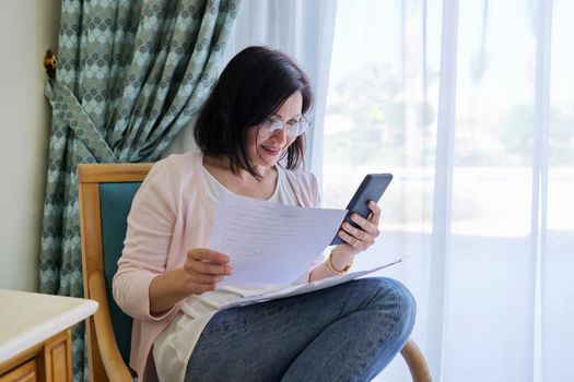 Middle age surprised woman with smartphone business papers sitting at home